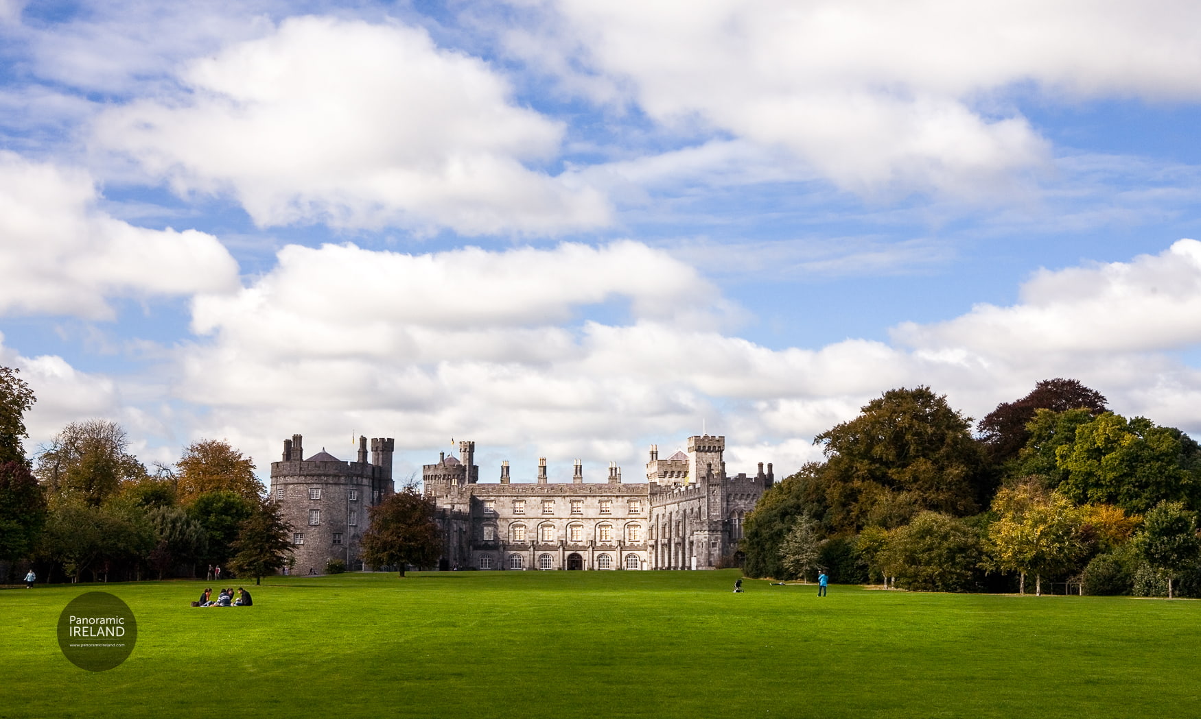 The grounds of Kilkenny Castle in Ireland on a blue sky day with green lawns, green trees, white clouds and people relaxing. This photo by Panoramic Ireland.