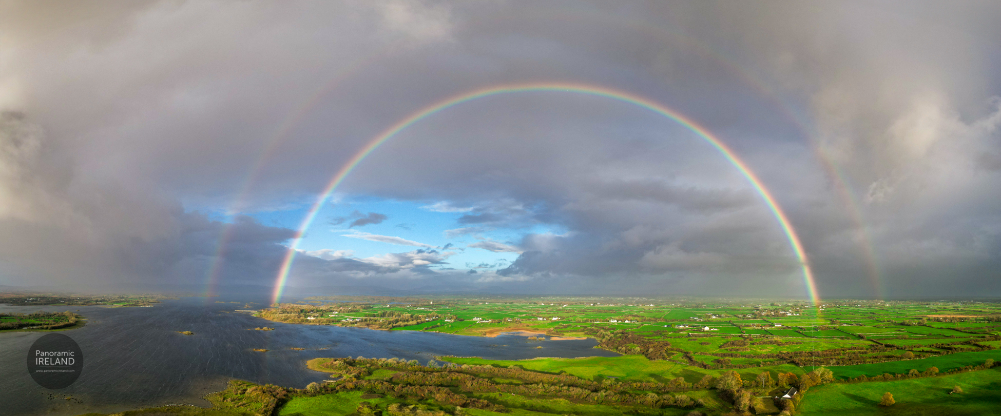 A sweeping panoramic, aerial view of a spectacular double rainbow arching over a vast Irish landscape. The primary rainbow is bright and forms a complete, perfect arc, with a fainter secondary rainbow visible above it. Beneath the rainbows, the land is a vibrant patchwork of bright green fields, dotted with small houses, trees, and stone walls. To the left, a large, dark body of water, likely a lough or wide river, reflects the sky. The weather is dramatic: dark, heavy storm clouds frame the scene on both sides, while a patch of bright blue sky with white clouds shines through directly under the rainbow's peak, indicating the sun is breaking through. Image by Panoramic Ireland.