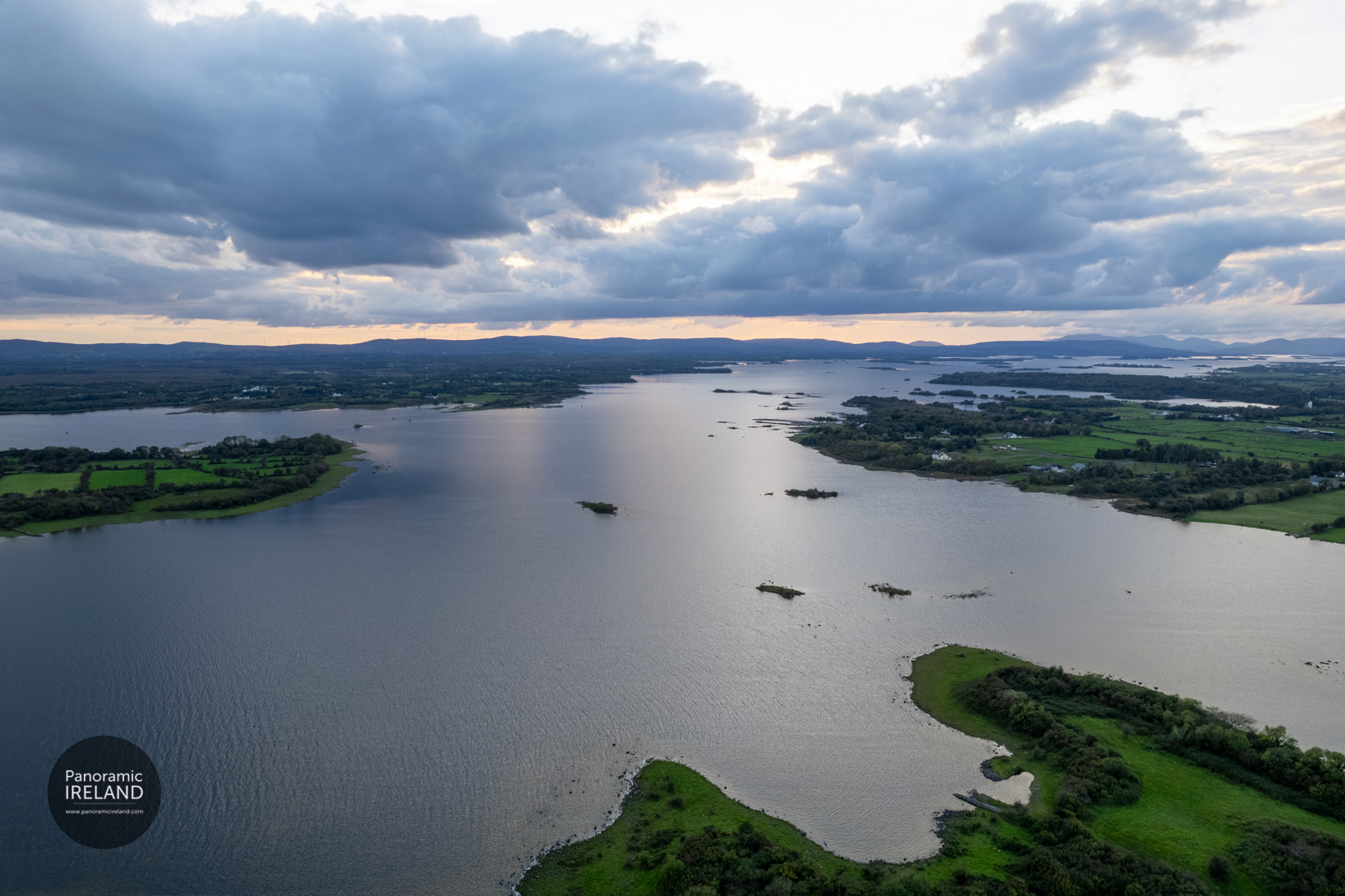 An expansive, aerial panoramic view of a large, tranquil lake in West Ireland, likely Lough Corrib or Lough Mask. Lush green peninsulas and small, tree-covered islands dot the dark, calm water. In the distance, rolling hills sit on the horizon under a dramatic, cloudy sky, with a soft glow from the setting sun breaking through the clouds at the horizon.