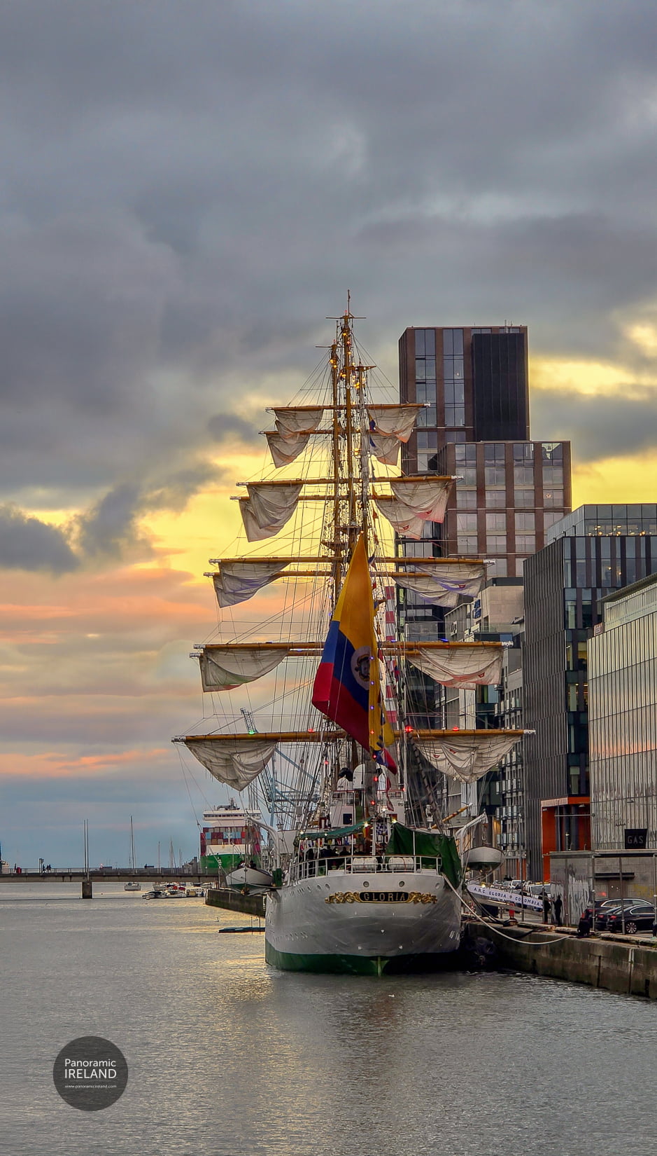 Gloria's stern and nameplate, the Colombian flag flying, the yards hung with furled sails against modern Dublin Docklands buildings. The stern of ARC Gloria with the Colombian flag, Dublin office buildings behind