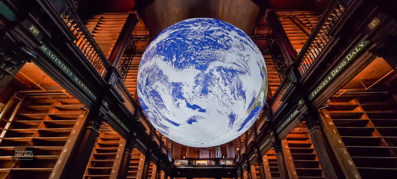Gaia floating beneath the vaulted wooden ceiling of Trinity College Dublin’s Long Room, with marble busts and empty upper shelves