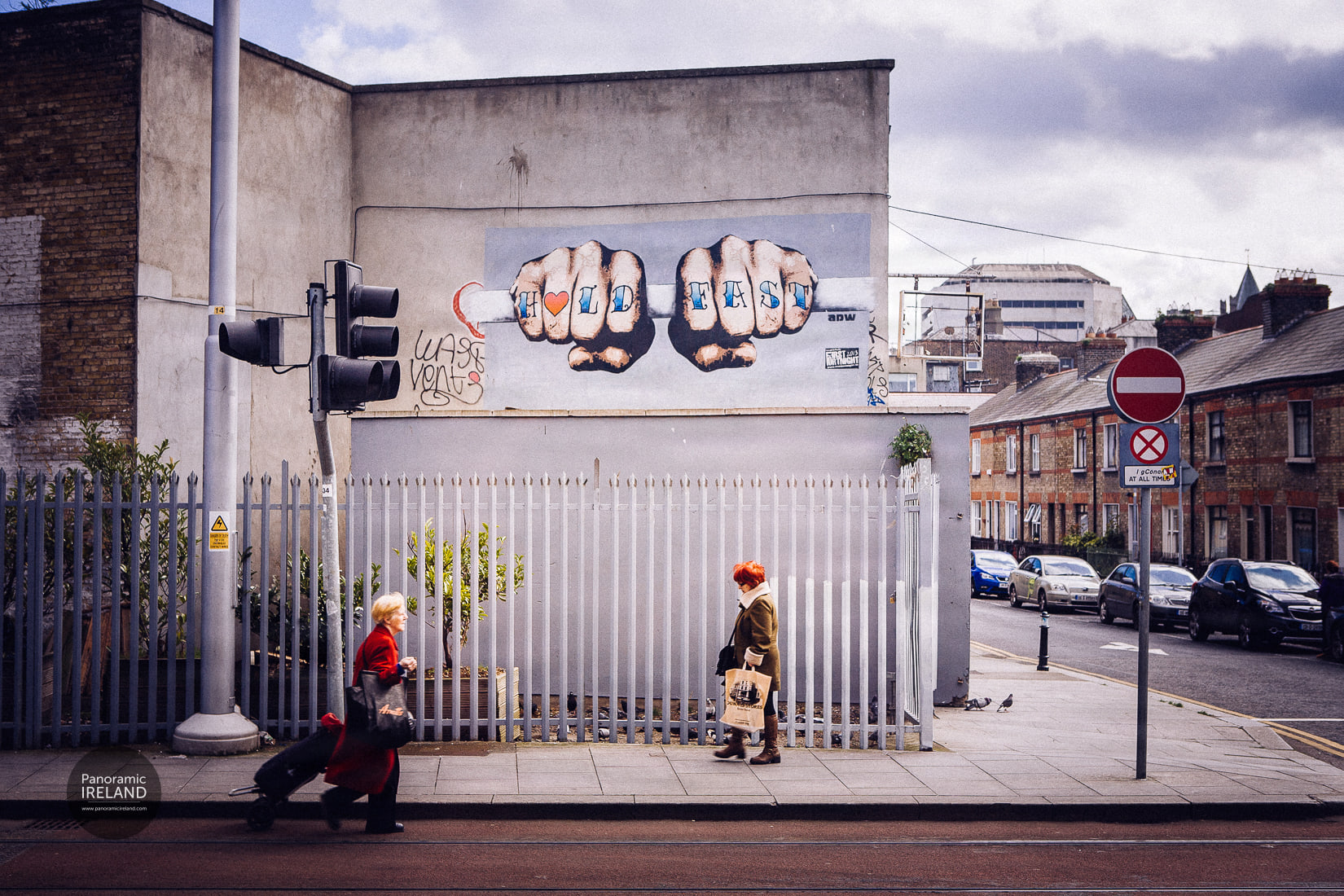 HOLD FAST street art mural by ADW in Dublin — two fists gripping a bar with HOLD FAST lettered across the knuckles, pedestrians passing on the pavement.