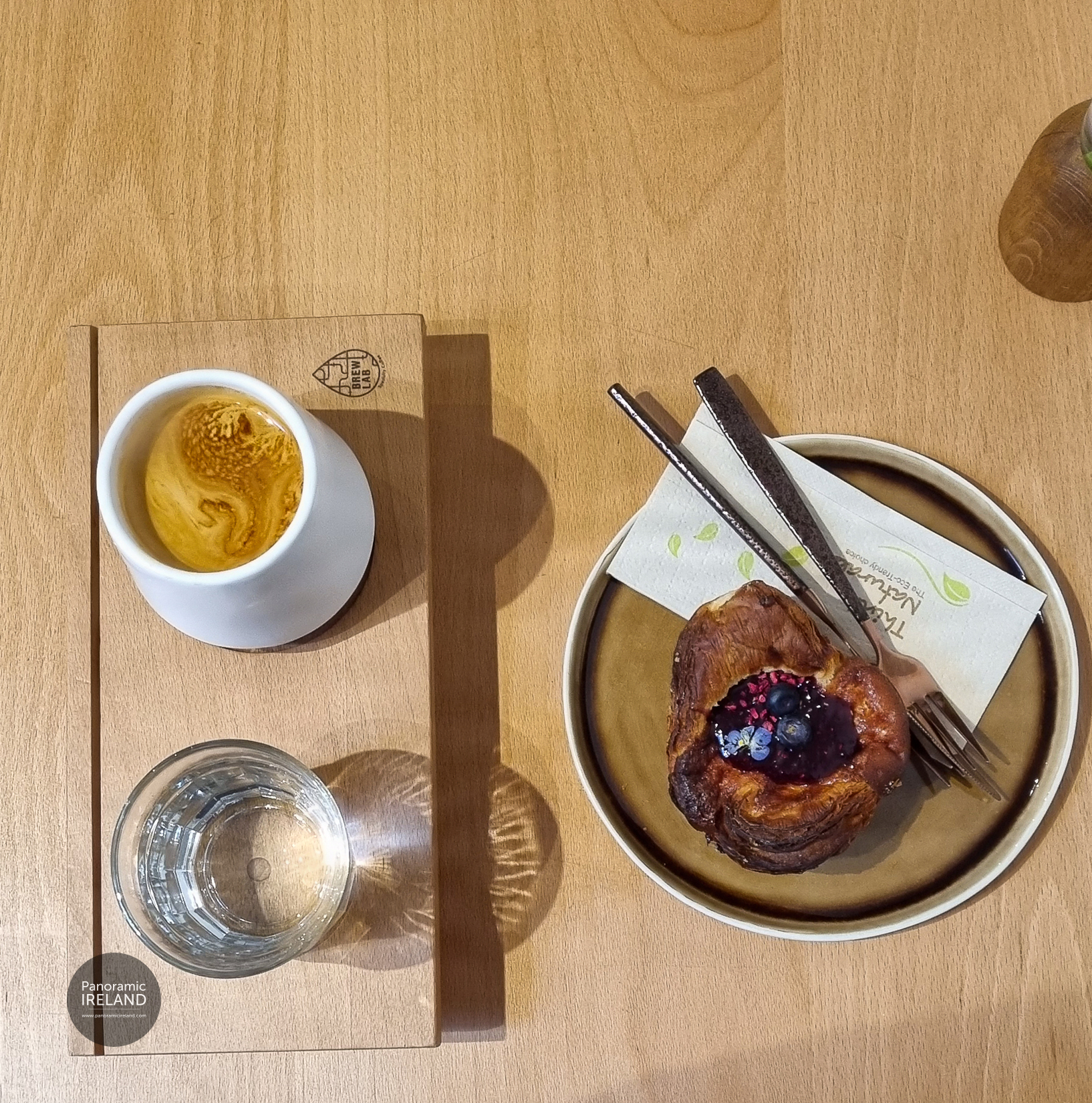 An americano in a thick-walled, matte-glazed stoneware cup, served on a Brew Lab wooden board with a glass of water. Beside it, a flaky pastry topped with berry compote, blueberries and a viola sits on a caramel-glazed stoneware plate. Everything is laid out on warm beech in low, raking light.
